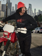 Alpinestars Pulse Hoodie, zwarte fleece pullover met capuchon met rood en grijs Astars Combo Logo, gedragen door een man met een oranje muts naast een rode Honda motorfiets in een stedelijke omgeving, casual motorsport streetwear