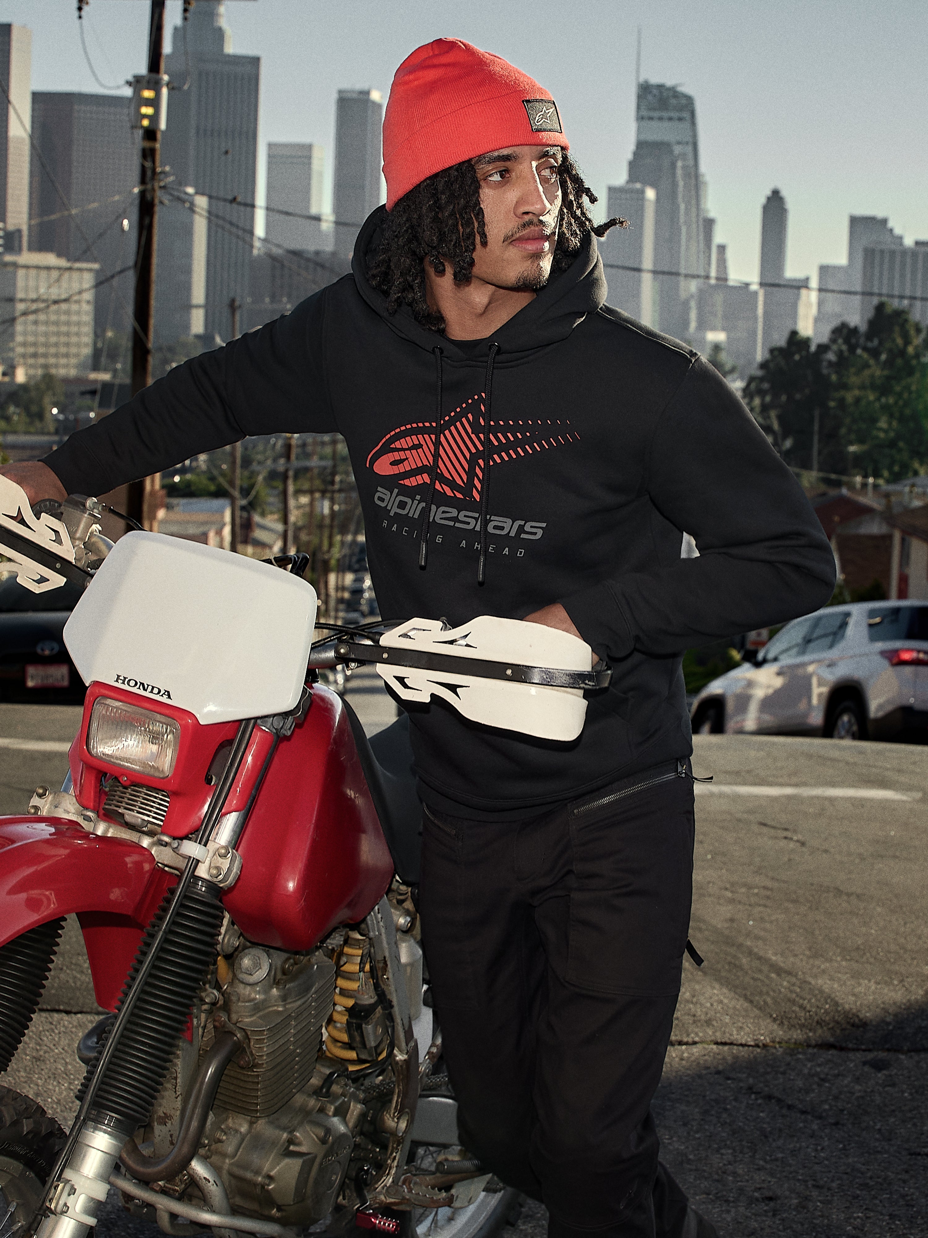 Alpinestars Pulse Hoodie, zwarte fleece pullover met capuchon met rood en grijs Astars Combo Logo, gedragen door een man met een oranje muts naast een rode Honda motorfiets in een stedelijke omgeving, casual motorsport streetwear