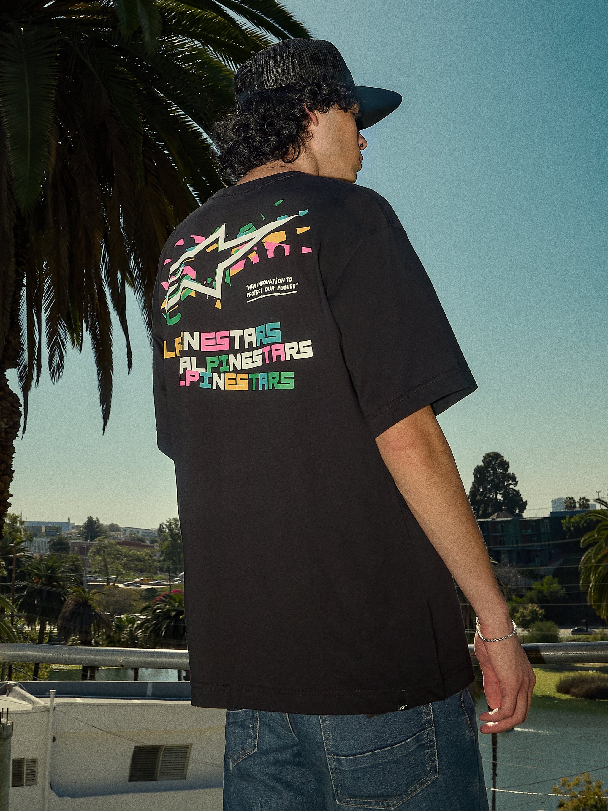 Alpinestars Wild Oversized CSF Tee, t-shirt à manches courtes noir, logo Astars multicolore éclatant au dos, coupe surdimensionnée, photo lifestyle streetwear décontractée avec des palmiers