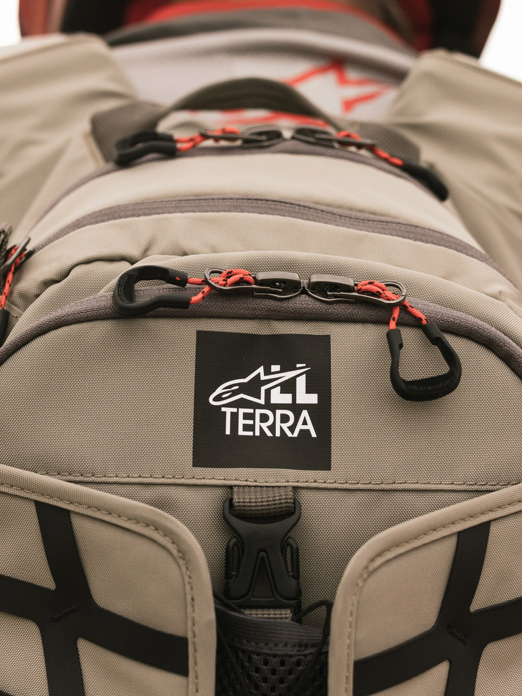 Alpinestars AT-15 Rucksack, Aluminum Walnut, Nahaufnahme des All Terra Logo-Patches auf beigem technischem Textil, mit orangefarbenen Kordel-Reißverschluss-Zügen und robusten Konstruktionsdetails