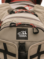 Alpinestars AT-15 Rucksack, Aluminum Walnut, Nahaufnahme des All Terra Logo-Patches auf beigem technischem Textil, mit orangefarbenen Kordel-Reißverschluss-Zügen und robusten Konstruktionsdetails