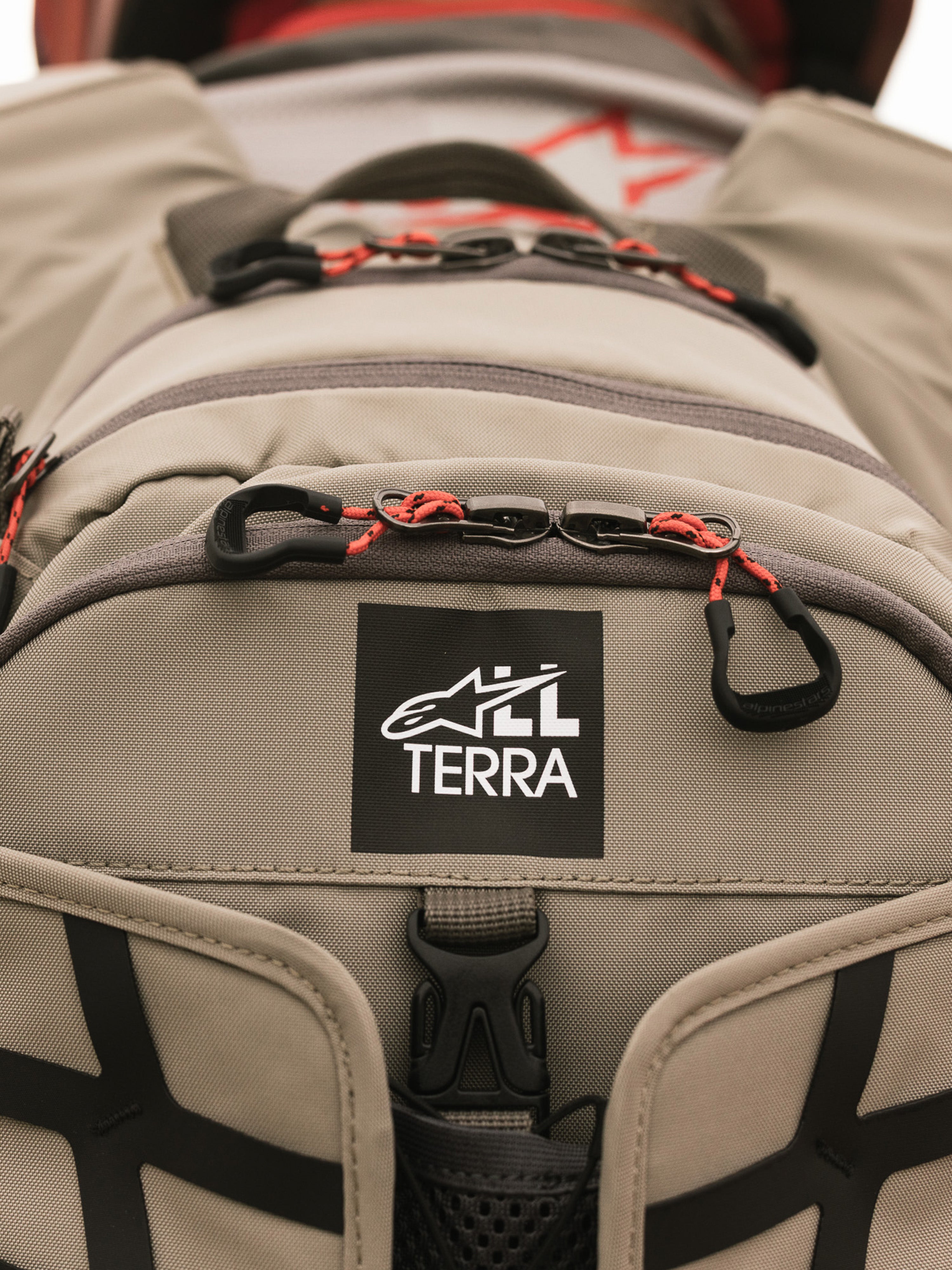 Alpinestars AT-15 Rucksack, Aluminum Walnut, Nahaufnahme des All Terra Logo-Patches auf beigem technischem Textil, mit orangefarbenen Kordel-Reißverschluss-Zügen und robusten Konstruktionsdetails