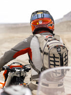 Alpinestars AT-15 Rucksack in Aluminum Walnut, getragen von einem Adventure-Motorradfahrer in einer Wüstenlandschaft. Robuste beige Textilkonstruktion mit schwarzem Gurtband, orangefarbenen Reißverschluss-Akzenten und 'All Terra'-Branding. Kombiniert mit einem rot-orangefarbenen Helm und grauer Fahrbekleidung für Offroad-Touren.