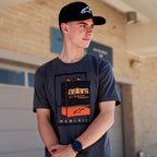 T-shirt Alpinestars Galun CSF, t-shirt à manches courtes, gris, motif graphique de bidon d'essence vintage à l'avant avec marquage « astars » et chiffres romains « MCMLXIII », porté par un homme avec une casquette noire à logo, style motorsport décontracté