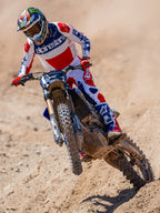 Motocross-Fahrer in Aktion mit Alpinestars Limited Edition Fortitude Tech 10 Stiefeln, Renn-Trikot und -Hose in hellem Rot, Weiß und Blau, abgestimmtes patriotisches Ausrüstungsset für Offroad-Performance auf einer Erdpiste