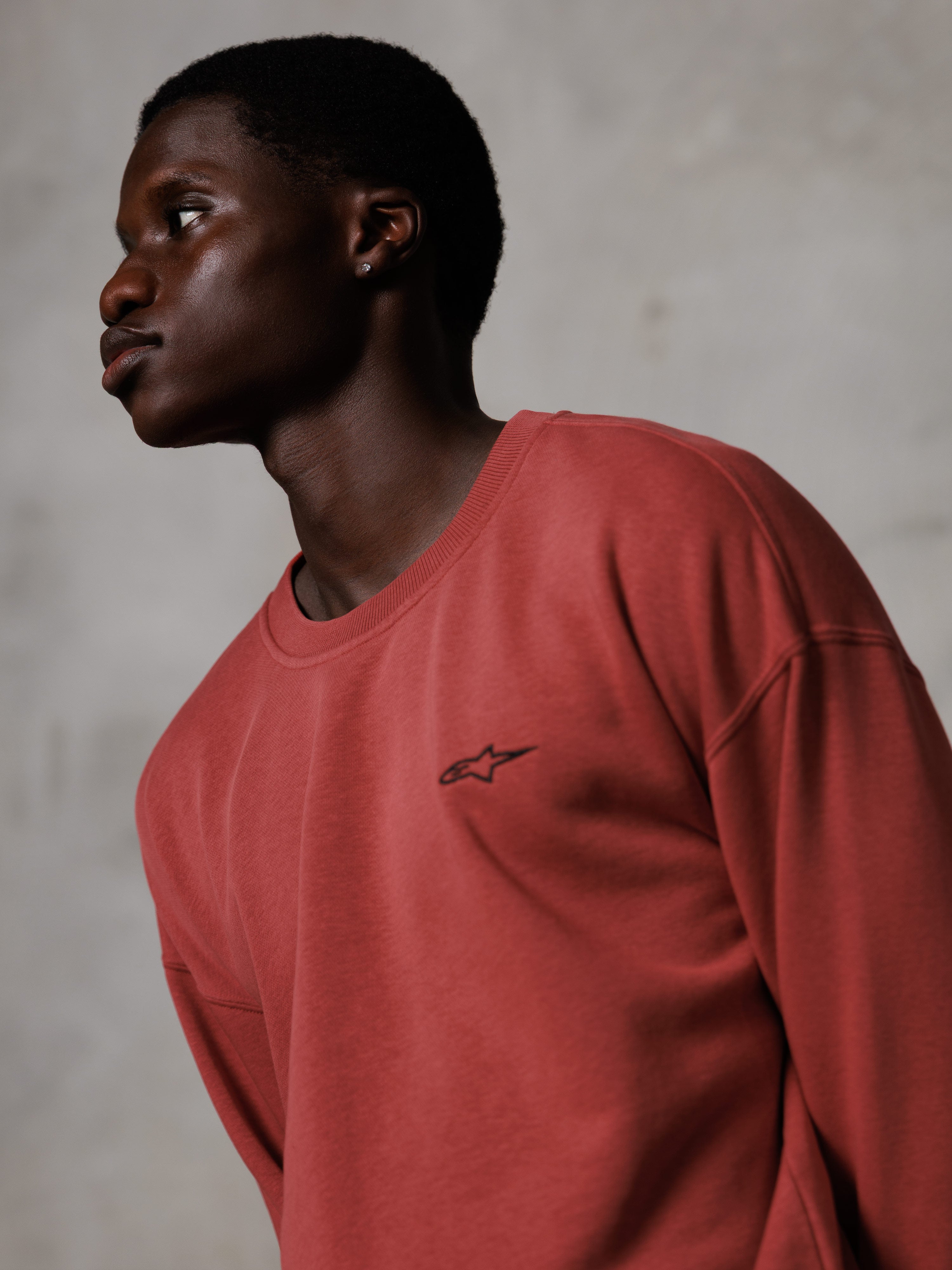 Alpinestars Essentials Ageless Crew, sweat-shirt à col rond rouge brique, porté par un mannequin homme de profil, petit logo étoile noir sur la poitrine, coupe décontractée, polaire premium, streetwear décontracté