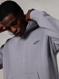 Alpinestars Essentials Ageless Hoodie, Silber, Nahaufnahme eines Mannes, der die Kapuze des grauen Sweatshirts richtet, kleines schwarzes Alpinestars-Logo auf der Brust, hochwertige, vom Motorsport inspirierte Freizeitkleidung