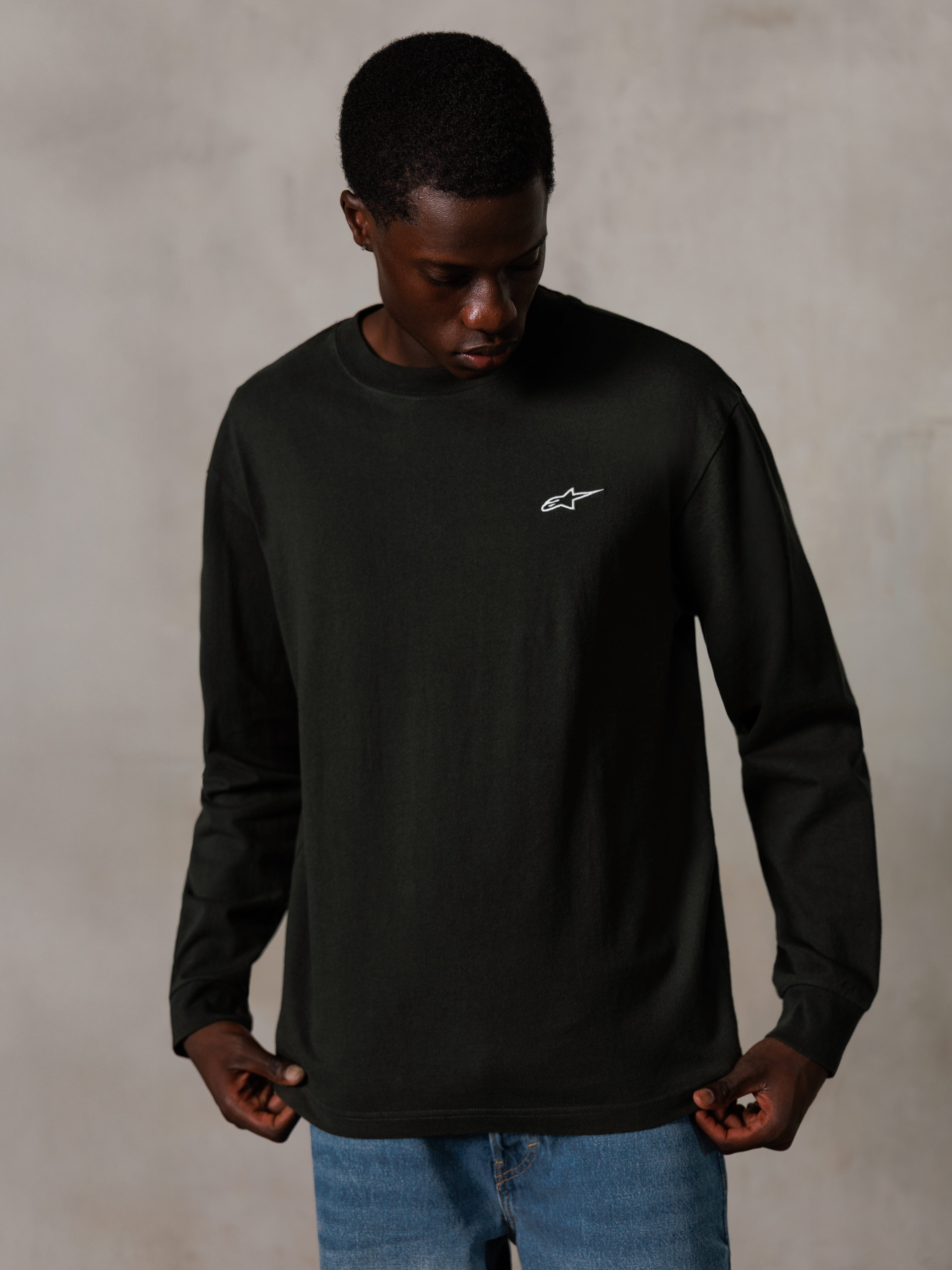 Alpinestars Essentials Ageless LS Tee, T-shirt met lange mouwen, Off Black, voorzien van een klein wit logo op de borst, gedragen door een model met blauwe jeans, casual lifestyle streetwear