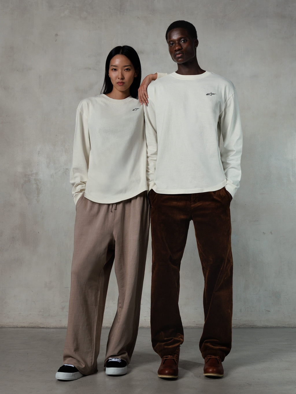 Alpinestars Essentials Ageless LS Tee, t-shirt à manches longues, blanc cassé, porté par des modèles homme et femme, petit logo Alpinestars noir sur la poitrine, coupe décontractée, associé à un pantalon de jogging beige et un pantalon en velours côtelé marron, style streetwear décontracté inspiré du sport automobile