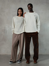 Alpinestars Essentials Ageless LS Tee, t-shirt à manches longues, blanc cassé, porté par des modèles homme et femme, petit logo Alpinestars noir sur la poitrine, coupe décontractée, associé à un pantalon de jogging beige et un pantalon en velours côtelé marron, style streetwear décontracté inspiré du sport automobile