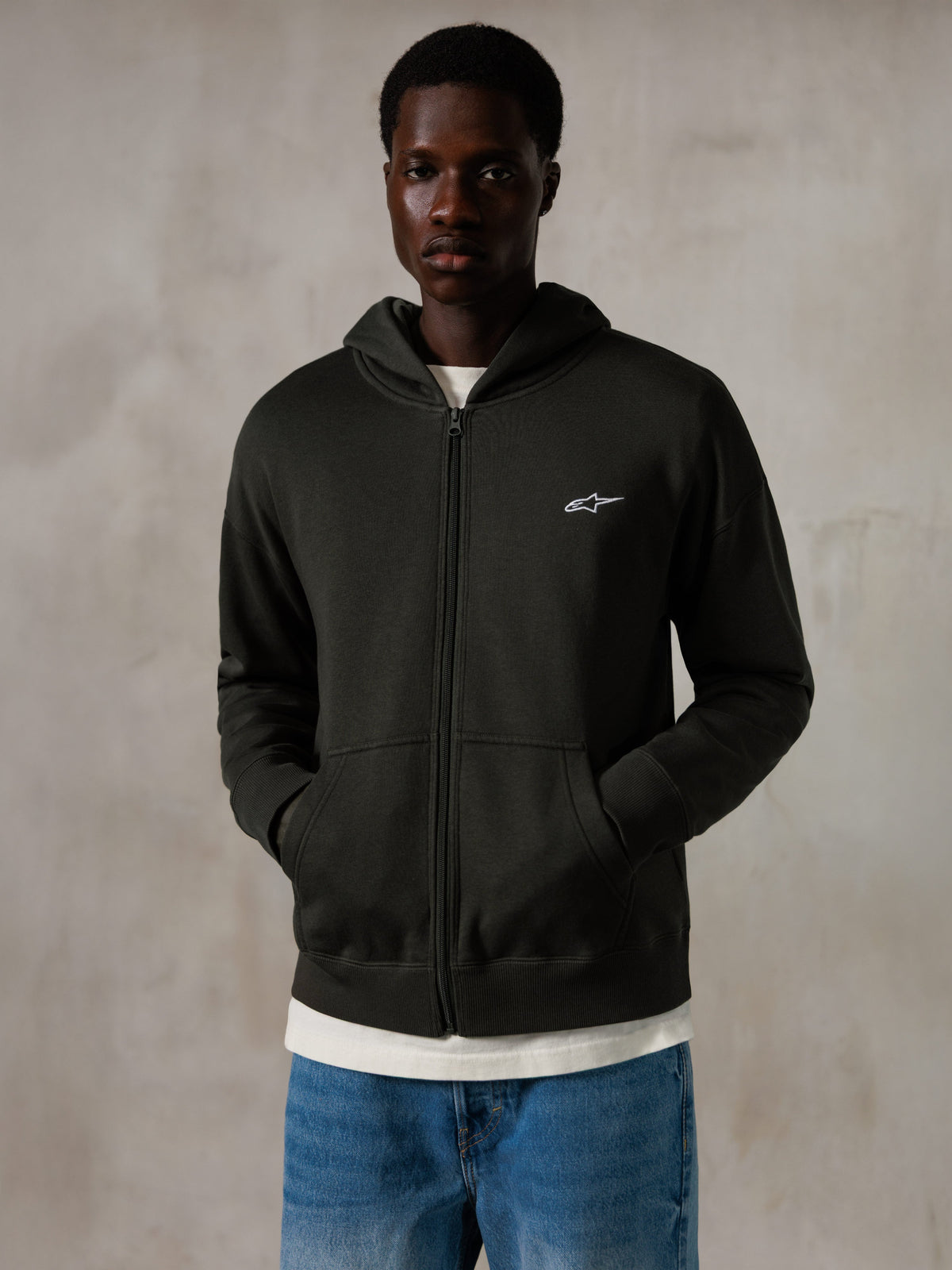 Sweat à capuche zippé Alpinestars Essentials Ageless, Off Black, sweat à capuche entièrement zippé avec un petit logo Alpinestars blanc sur la poitrine, coupe décontractée, porté par un mannequin avec un jean bleu et un t-shirt blanc, style streetwear décontracté inspiré du sport automobile