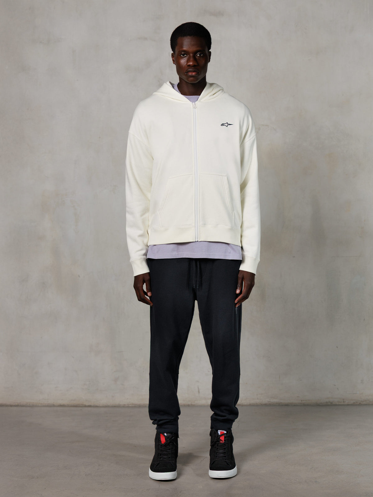 Sweat à capuche zippé Alpinestars Essentials Ageless, style streetwear décontracté, blanc cassé avec logo noir discret sur la poitrine, fermeture éclair intégrale, design à capuche, coupe décontractée, porté avec un pantalon de jogging noir et des baskets