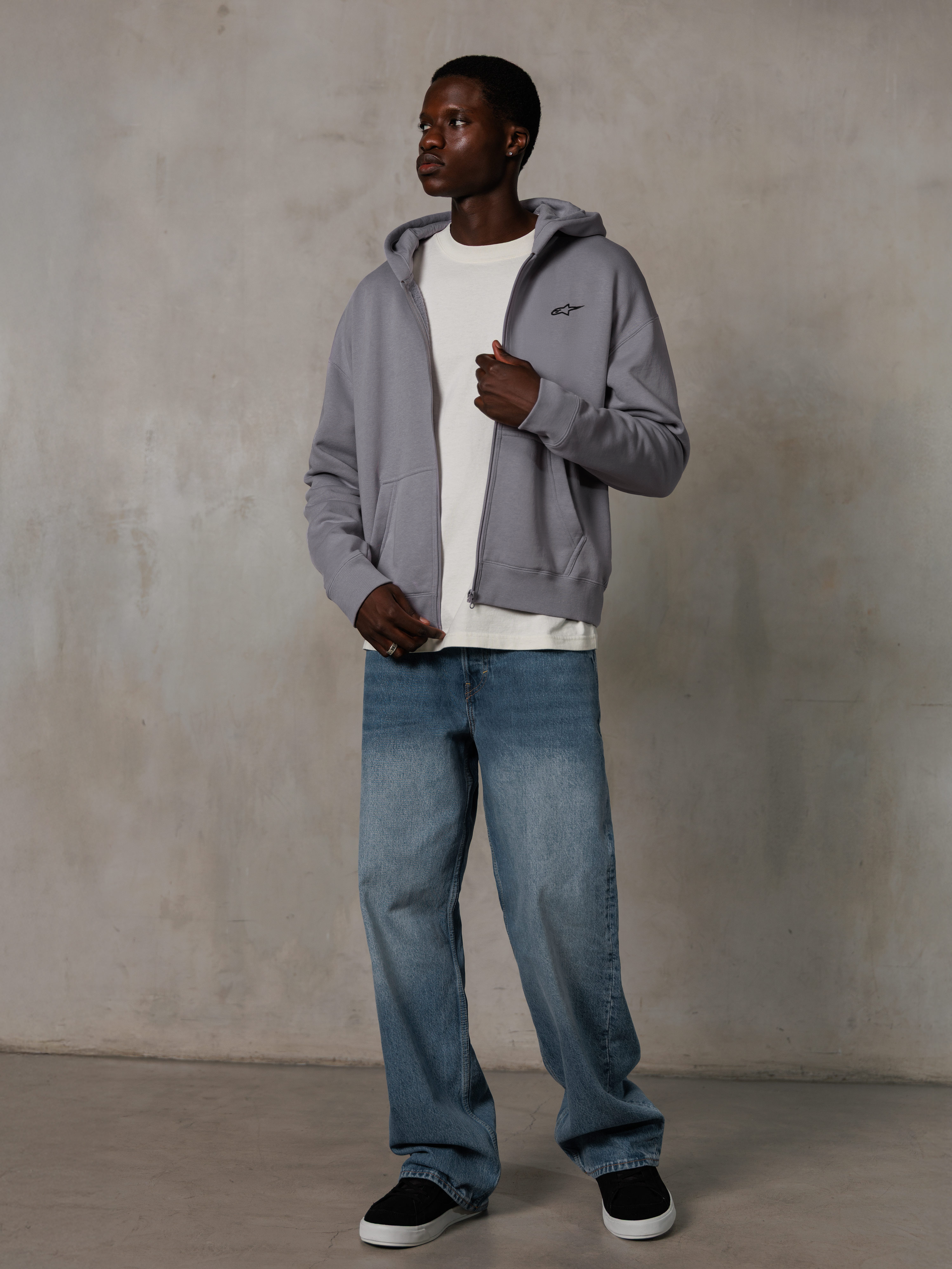 Alpinestars Essentials Ageless Zip Hoodie, Silver, streetwear décontracté, sweat à capuche zippé en polaire avec un petit logo noir sur la poitrine, porté par un mannequin avec un t-shirt blanc et un jean bleu, coupe lifestyle décontractée