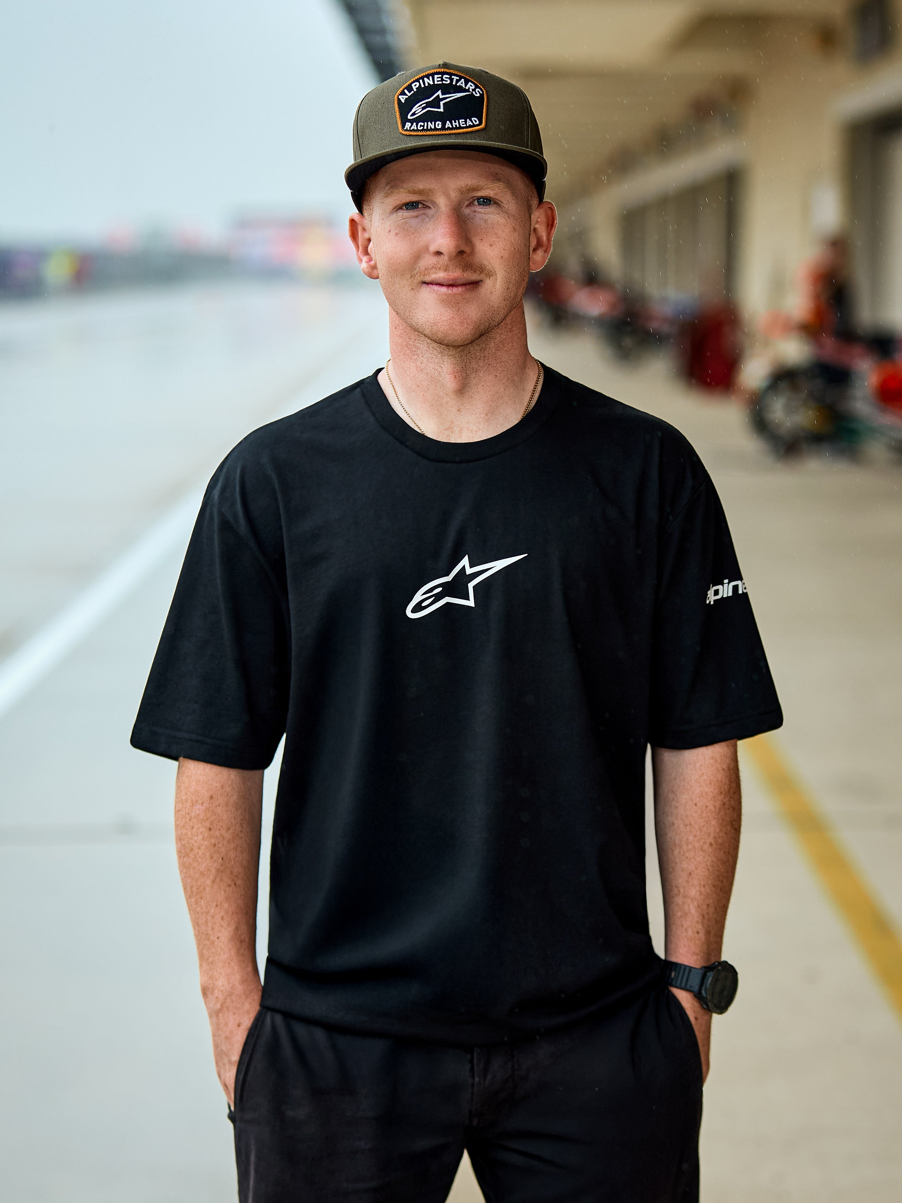 Man in Alpinestars Frontal CSF Tee, T-shirt met korte mouwen, zwart, wit centraal Astars-logo, olijfgroene Alpinestars pet, casual op motorsport geïnspireerde streetwear