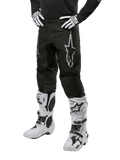 Alpinestars 2024 Fluid Graphite Pants, pantalon de motocross, noir et argent, avec un logo Alpinestars argenté sur la cuisse, présenté porté avec des bottes et des gants de motocross blancs, conçu pour la course de moto tout-terrain