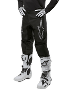 Alpinestars 2024 Fluid Graphite Pants, pantalon de motocross, noir et argent, avec un logo Alpinestars argenté sur la cuisse, présenté porté avec des bottes et des gants de motocross blancs, conçu pour la course de moto tout-terrain