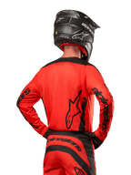 Alpinestars 2024 Fluid Lurv Jersey, motorcross shirt van achteren gezien, Hot Orange en zwart, opvallende Alpinestars-branding op de rug en mouwen, bijpassende crossbroek en zwarte helm, ontworpen voor off-road raceprestaties