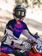 Motocross-Fahrer auf einem roten Motocross-Bike, Limited Edition HUNTA96 XXV Supertech Vision Schutzbrille in Lila und Weiß mit silbernen Spiegelgläsern, passendes lila-schwarz-weißes Alpinestars-Jersey und Hose, Red Bull-Helm, weiße Handschuhe, koordinierte professionelle Motocross-Rennausrüstung.