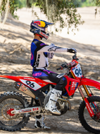 Motocross-Fahrer mit der Limited Edition HUNTA96 XXV Supertech Vision Brille in Lila, Weiß und Silber, passendes lila-weißes Renntrikot mit weißen Stiefeln, sitzend auf einer roten Honda Motocross-Maschine mit der Startnummer 96, Outdoor-Offroad-Einsatz auf einer Sandstrecke.