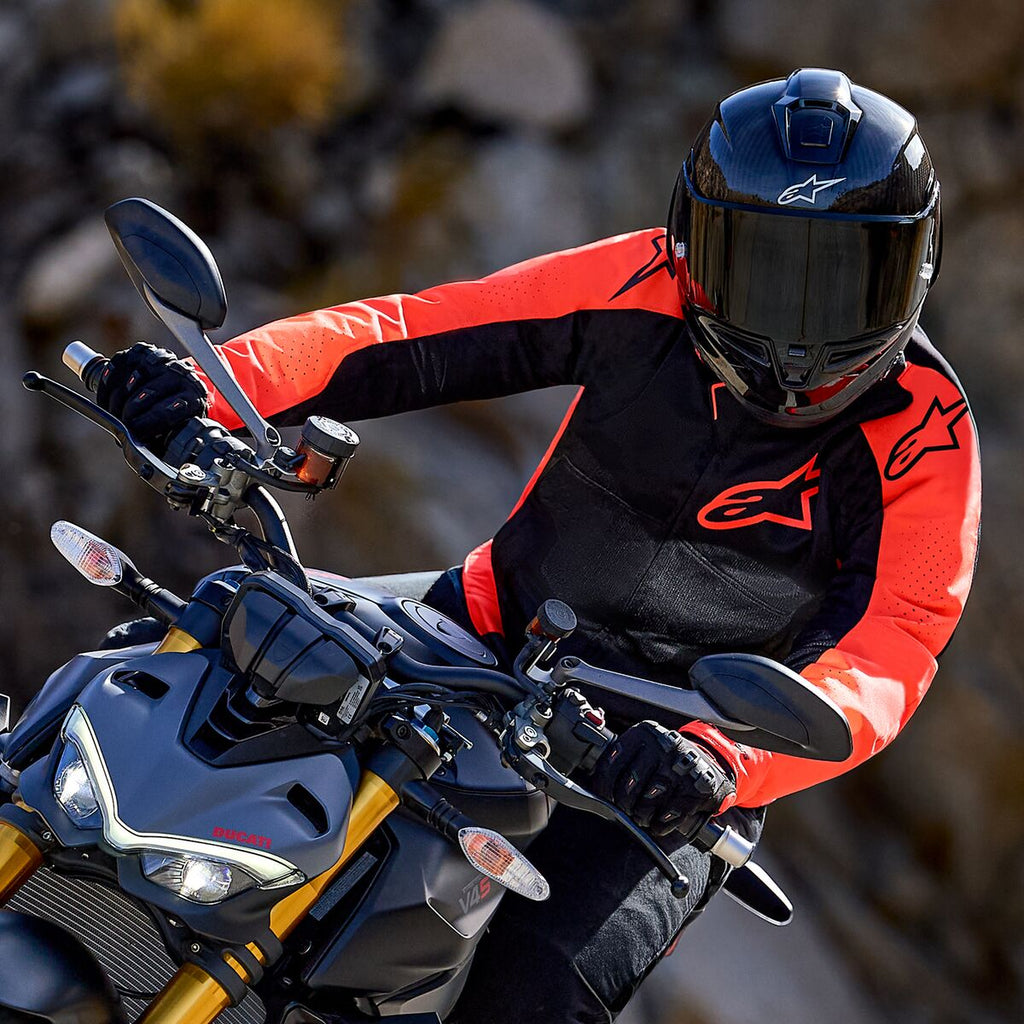 Veste Alpinestars Viper Air V4, rouge fluo et noir, portée par un motard penché dans un virage sur une moto sportive, avec casque intégral noir et gants, équipement de course et de sport haute performance
