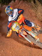 Motocross rider leaning into a turn on an orange motorcycle, wearing Alpinestars Pro-Dura Jersey and Pants in Navy, Hot Orange, and White, coordinated off-road racing gear, Red Bull branded helmet, white boots, action shot on a dusty dirt track.