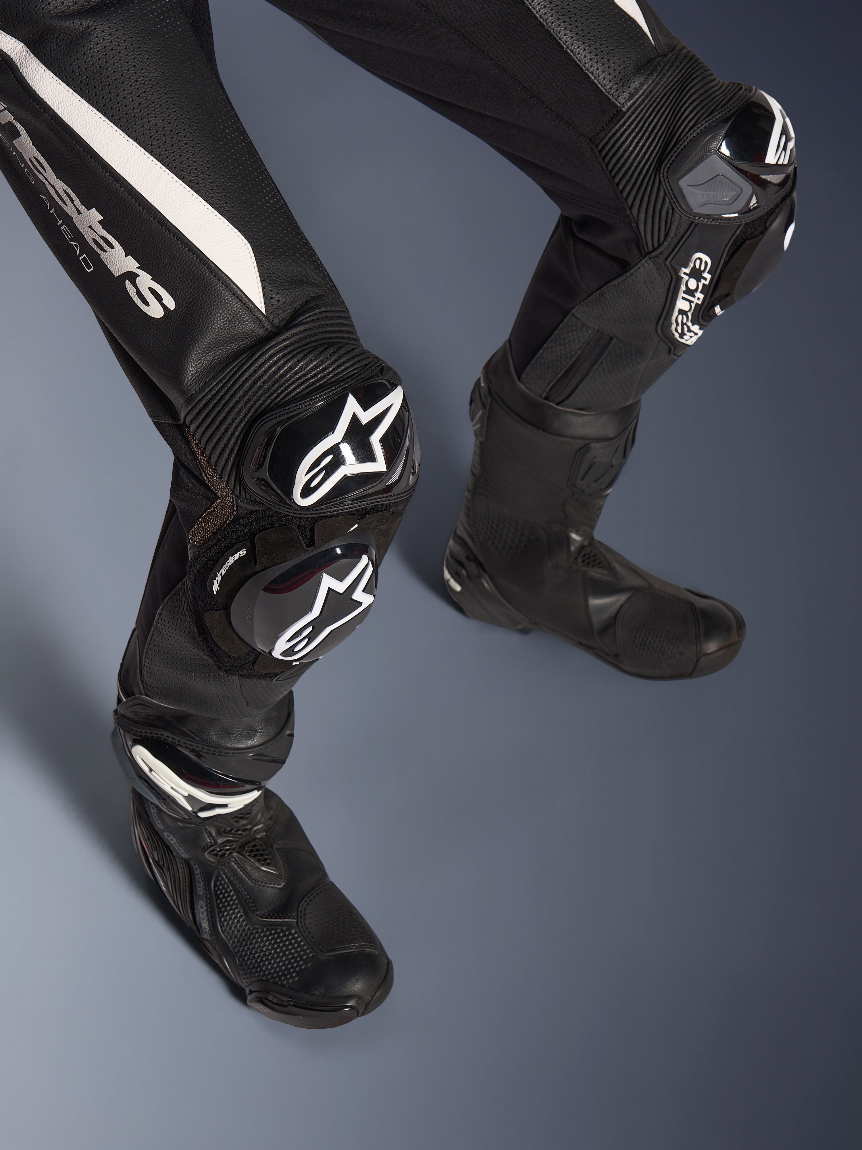Detail van de benen van het Alpinestars Stella GP-R7 2-delig leren pak, zwart met witte accenten, voorzien van GP DFS RR kniebescherming en sliders met Alpinestars-branding, gecombineerd met zwarte racelaarzen, ontworpen voor circuitprestaties