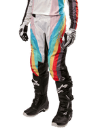 Alpinestars Stella Techstar 2024, pantalon de motocross pour femme, noir multicolore avec rayures diagonales rouges, jaunes et bleues, porté avec des bottes de moto noires et des gants blancs, avec panneaux de protection noirs aux genoux et marquage audacieux pour la course tout-terrain