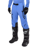 Alpinestars 2024 Supertech Dade Pants, pantalon de motocross, bleu clair avec logos graphiques noirs, avec perforations de ventilation et boucle magnétique à la taille, porté avec des bottes de pilotage techniques et des gants noirs, équipement de course tout-terrain professionnel