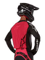 Alpinestars 2024 Supertech Ward Trikot, Motocross-Trikot, Schwarz und Red Berry, Rückansicht mit rotem Mesh-Einsatz und schwarzen Ärmeln mit weißem Branding, getragen mit schwarzem Carbon-Helm und Handschuhen, athletische Rennpassform für den Offroad-Einsatz