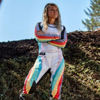 Pilote de motocross femme portant le pantalon Alpinestars Stella Techstar 2024 et le maillot assorti, design Noir Multicolore avec base blanche et rayures éclatantes bleues, jaunes et rouges, debout les bras croisés sur une piste de terre, tenue de course tout-terrain professionnelle