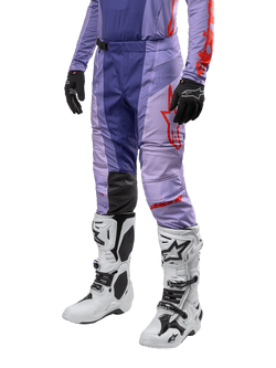 Alpinestars 2024 Techstar Ocuri crossbroek, motorcross broek in paars met fel oranje en rode accenten, rood logo op het bovenbeen, gedragen met witte motorcrosslaarzen en zwarte handschoenen, duurzame stretchconstructie voor off-road gebruik.