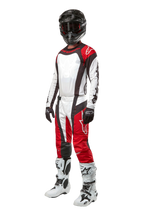 Pilote de motocross portant une tenue complète Alpinestars, pantalon Techstar Ocuri 2024 en rouge Mars, blanc et noir, maillot assorti, casque noir avec lunettes, gants noirs et blancs, et bottes de motocross blanches, tenue de course tout-terrain professionnelle