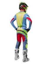 Motorcrosser van achteren gezien in 2024 Techstar Ocuri-broek en bijpassende jersey, lichtblauw, fluor geel en bessenrood, witte en zwarte laarzen, zwarte helm met neon accenten, gecoördineerde Alpinestars-uitrusting voor off-road racen