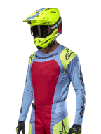 Alpinestars 2024 Techstar Ocuri Jersey, motorcross raceshirt, lichtblauw met rode en neongele accenten, gedragen met bijpassende neongele helm en zwarte crossbril, voorzien van zwarte Alpinestars-logo's op de borst en schouders, atletisch race-geïnspireerd ontwerp voor off-road prestaties