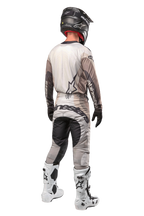Alpinestars 2024 Techstar Pneuma Pants, pantalon de motocross, sable foncé et gris, vue arrière porté par un pilote avec un maillot assorti, des bottes blanches et noires et un casque noir, avec un marquage Alpinestars imposant sur l'assise et des panneaux sombres renforcés