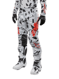 Alpinestars Pantalon Techstar Rantera 2024, pantalon de motocross, camouflage gris brume avec motifs abstraits noirs et rouges, porté avec un maillot assorti, des gants noirs et des bottes de motocross blanches, conçu pour la course tout-terrain haute performance