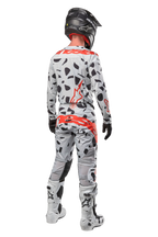 Pilote de motocross vu de dos portant le pantalon Alpinestars Techstar Rantera 2024, camouflage gris avec taches noires et logos rouges, maillot assorti, casque noir et bottes blanches, tenue de course tout-terrain complète