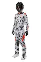 Pantalon Alpinestars Techstar Rantera 2024, pantalon de motocross, camouflage gris, porté avec un maillot assorti, un casque noir et des bottes blanches et noires, avec des motifs abstraits noirs et des accents rouges, équipement complet de course tout-terrain