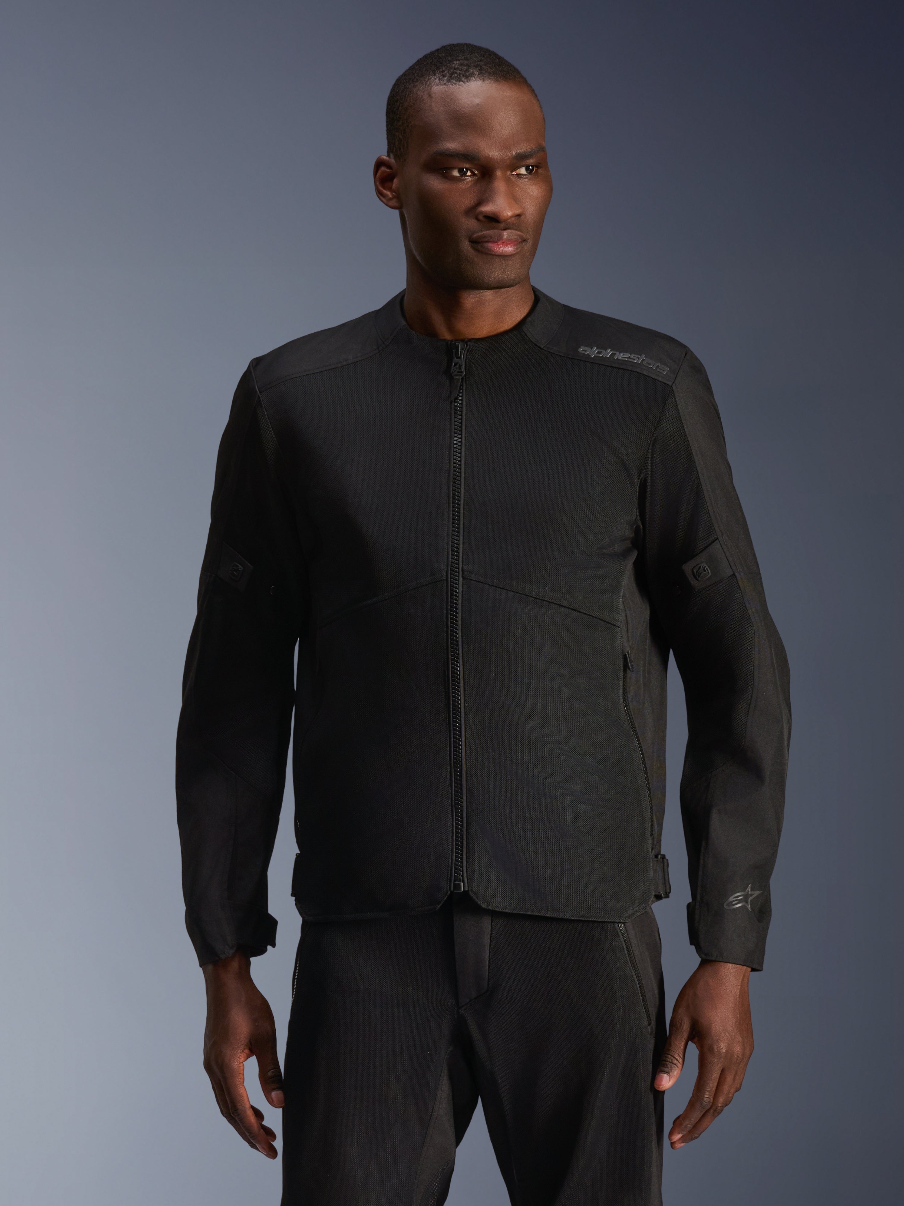 Mannequin homme portant la veste Alpinestars C-1 Air noire, une veste de moto contemporaine dotée de larges panneaux en maille technique et d'une fermeture éclair frontale intégrale, associée à un pantalon de moto noir assorti, esthétique urbaine et trajets quotidiens
