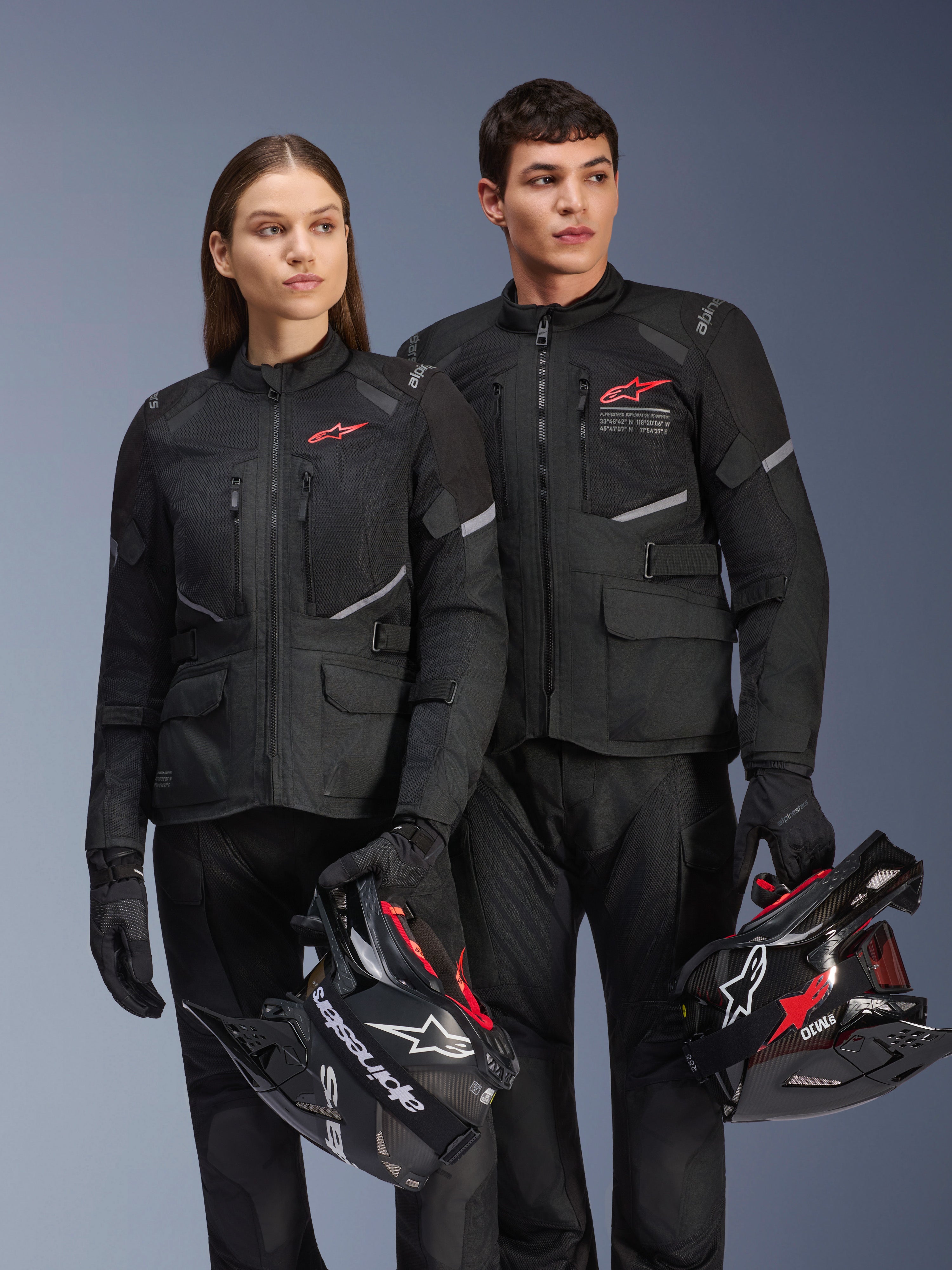 Man en vrouw dragen Alpinestars Andes Air Drystar®-jassen, zwarte adventure touring-kleding met mesh-panelen en reflecterende details, bijpassende zwarte broek en handschoenen, houden motorcrosshelmen vast, studio-opname van professionele motorkleding