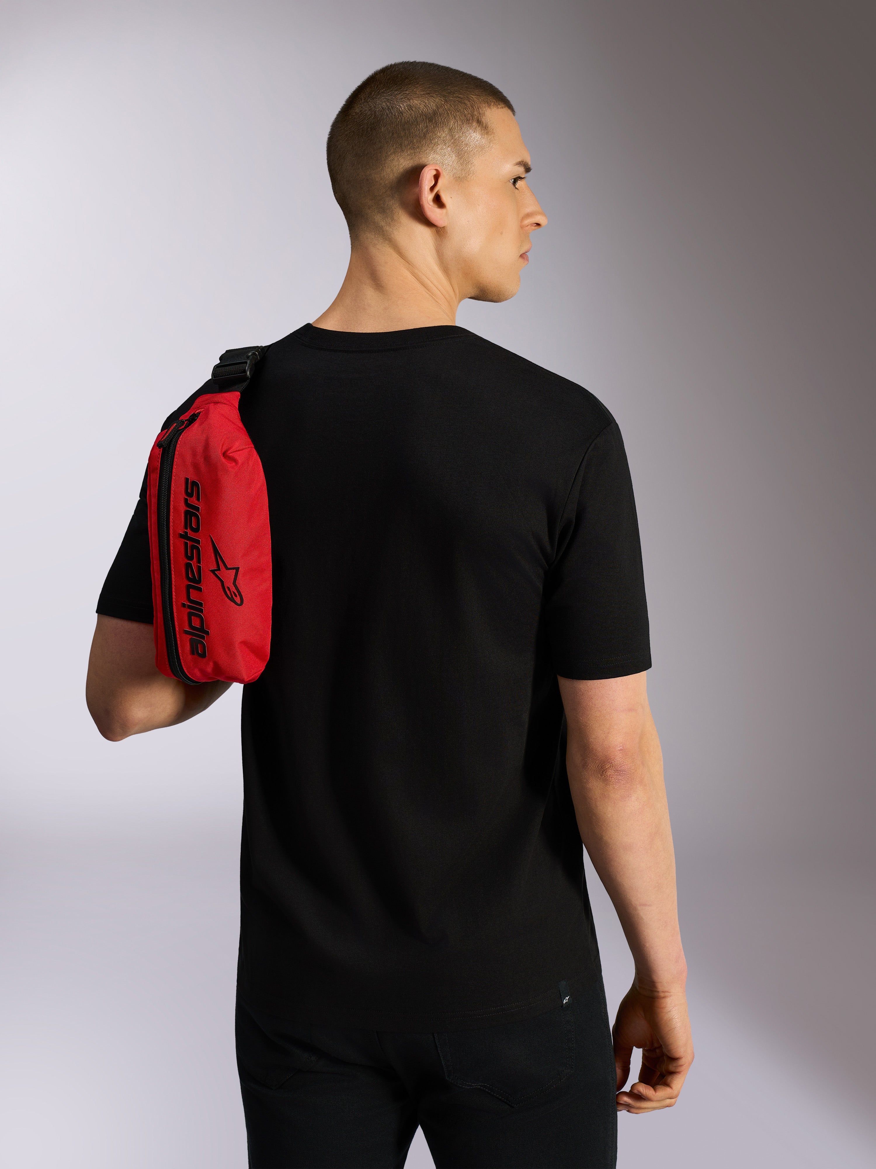 Alpinestars Only Linear heuptas, rood en zwart, over de schouder gedragen als slingbag door een model in een zwart t-shirt, voorzien van een verticale ritssluiting en zwarte Alpinestars-branding, duurzaam urban accessoire.